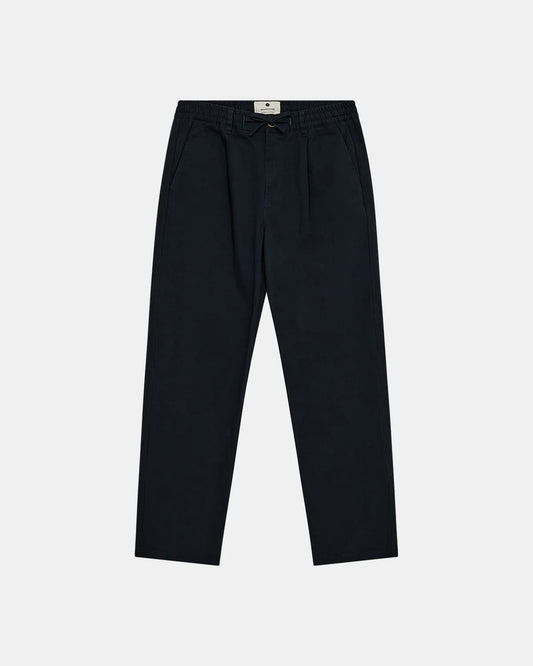 Vista frontale flat lay dei pantaloni Anerkjendt AKJAN Cotton Pleat Ela Pants in Navy Blue scuro su sfondo grigio chiaro: pantaloni uomo vita elasticizzata con coulisse laccetto nero doppia pinces frontale tasche laterali a filetto gamba tapered e patch etichetta rotonda Anerkjendt