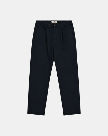 Vista frontale flat lay dei pantaloni Anerkjendt AKJAN Cotton Pleat Ela Pants in Navy Blue scuro su sfondo grigio chiaro: pantaloni uomo vita elasticizzata con coulisse laccetto nero doppia pinces frontale tasche laterali a filetto gamba tapered e patch etichetta rotonda Anerkjendt