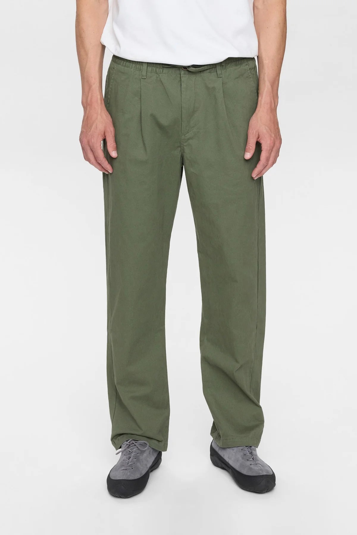 Vista frontale intera del modello che indossa i pantaloni Anerkjendt AKJAN Cotton Pleat Ela Pants in Khaki Sand con t-shirt panna e New Balance grigie: pantaloni uomo vita elastica doppia pinces frontale gamba tapered cotone beige sabbia su sfondo bianco