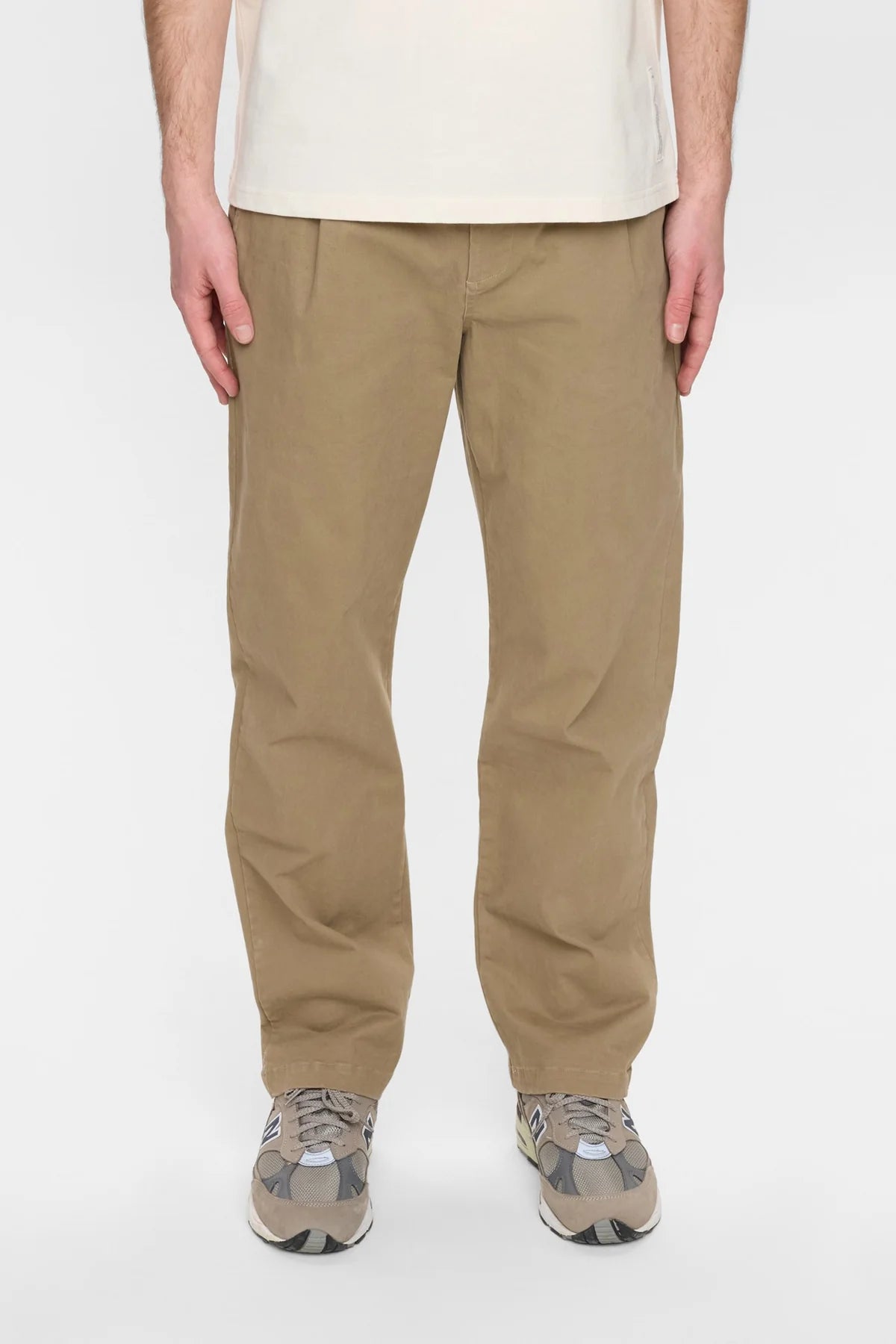 Vista frontale intera del modello che indossa i pantaloni Anerkjendt AKJAN Cotton Pleat Ela Pants con t-shirt e sneaker: pantaloni uomo vita elastica coulisse doppia pinces frontale gamba tapered su sfondo bianco