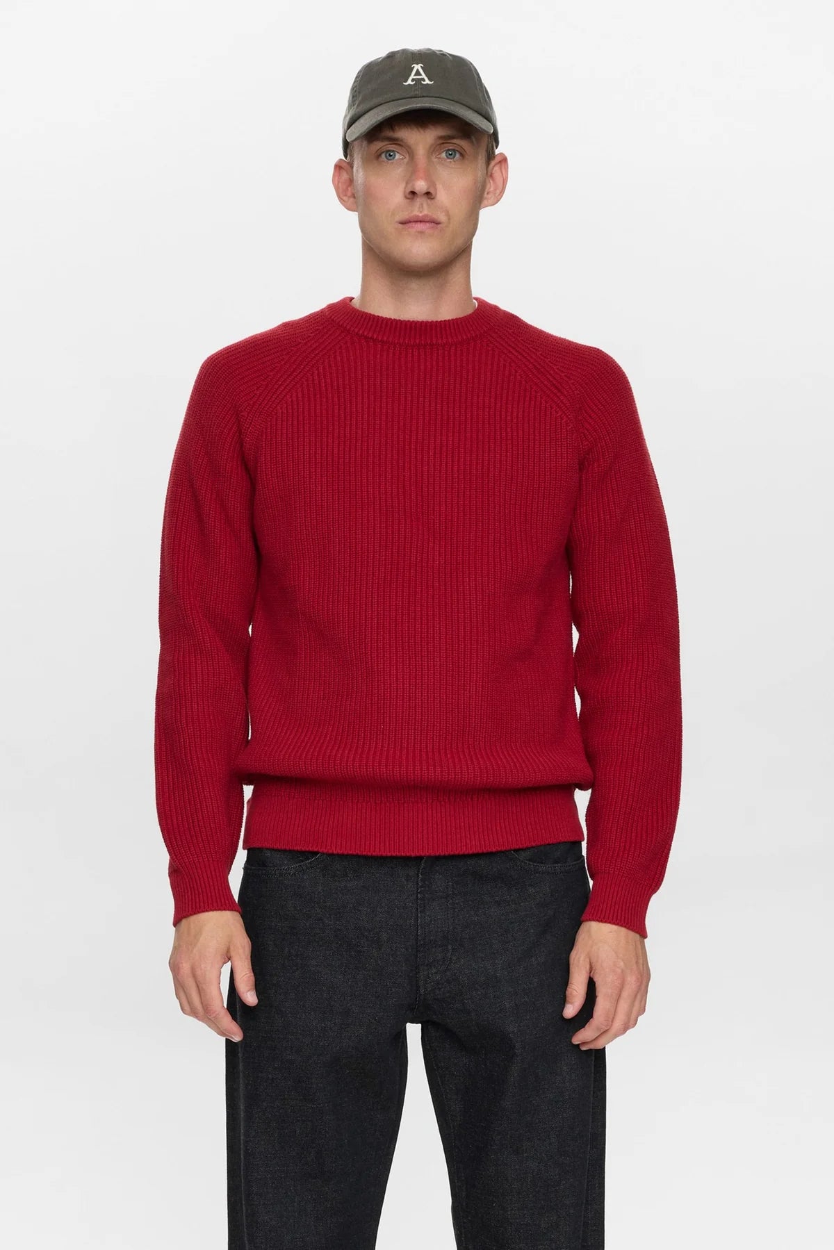 Vista posteriore intera del modello che indossa il maglione Anerkjendt AKSUNE Patent Knit rosso bordeaux con cappellino verde oliva pantaloni neri e scarpe nere: retro del maglione a costine con cuciture raglan diagonali e etichetta bianca Anerkjendt sul fondo