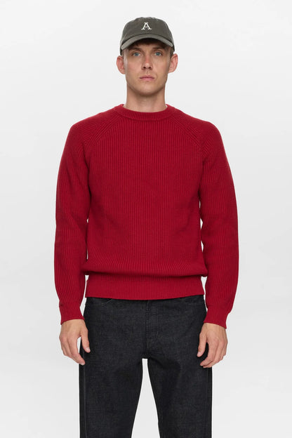 Vista posteriore intera del modello che indossa il maglione Anerkjendt AKSUNE Patent Knit rosso bordeaux con cappellino verde oliva pantaloni neri e scarpe nere: retro del maglione a costine con cuciture raglan diagonali e etichetta bianca Anerkjendt sul fondo