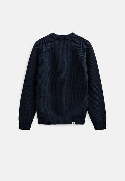 Modello maschile con cappellino verde oliva Anerkjendt indossa il maglione AKSUNE Patent Knit Navy Blue con jeans baggy lavaggio medio e sneaker grigie: vista frontale completa testa-piedi su sfondo bianco con maglione a costine blu navy e collo rotondo
