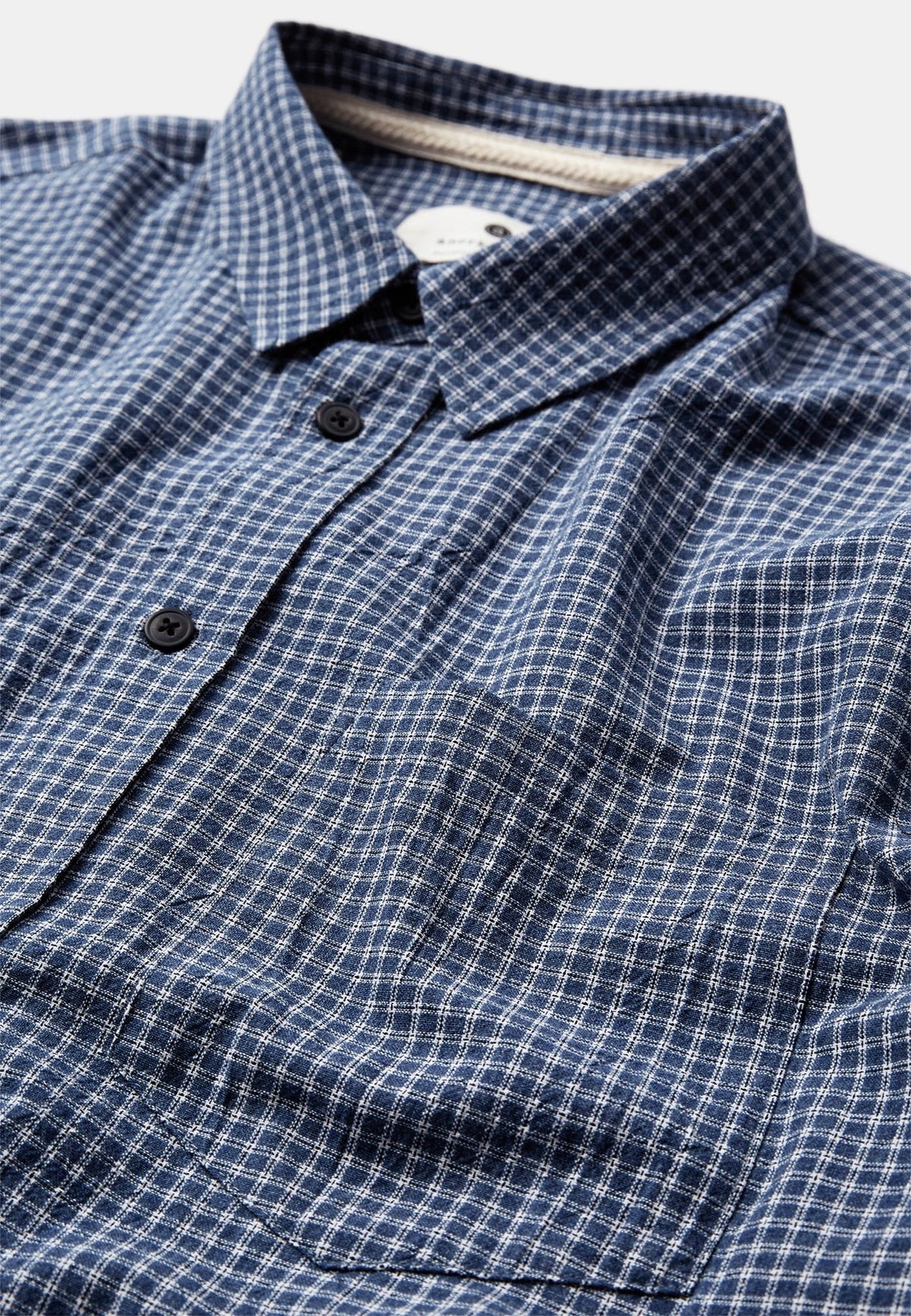 Dettaglio ravvicinato del colletto e della tasca a toppa sul petto della Anerkjendt AKOTTO Small Check LS Shirt Estate Blue: primo piano del tessuto a micro quadri blu navy e bianco con bottoni scuri tonali e colletto classico aperto su t-shirt bianca