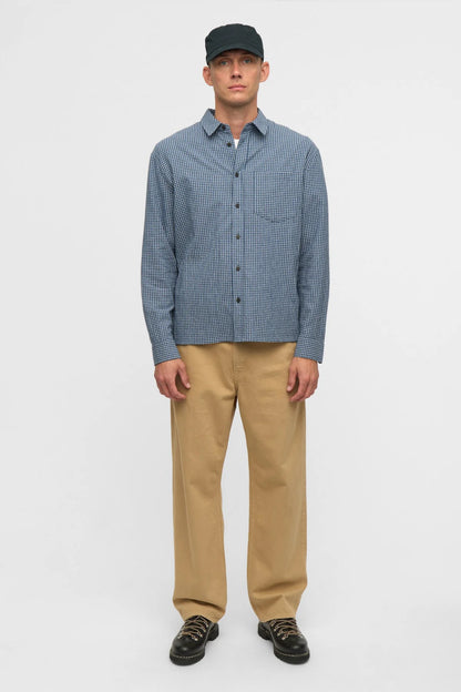 Vista posteriore del modello che indossa la Anerkjendt AKOTTO Small Check LS Shirt Estate Blue con cappellino nero pantaloni chino beige e scarpe nere: retro della camicia a micro quadri blu navy con yoke posteriore e piega centrale su sfondo bianco