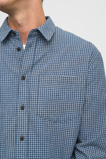 Vista frontale flat lay della Anerkjendt AKOTTO Small Check LS Shirt Estate Blue: camicia uomo a micro quadri blu navy e bianco con colletto classico, bottoni scuri, tasca a toppa sul petto e polsini a doppio bottone su sfondo grigio chiaro