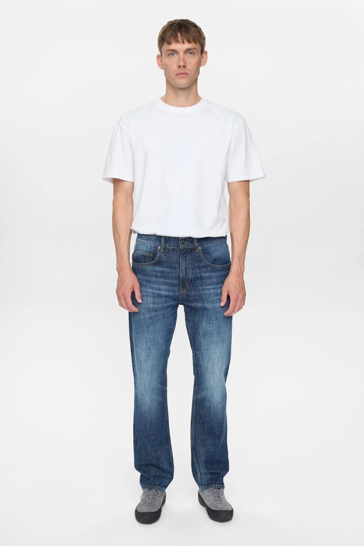 Vista laterale sinistra del modello che indossa i jeans Anerkjendt AKJUSTIN Dark Blue Sky con t-shirt bianca e sneaker grigie: profilo dei jeans straight fit che mostra la gamba dritta dal fianco all orlo grezzo con texture jacquard su denim blu medio su sfondo bianco