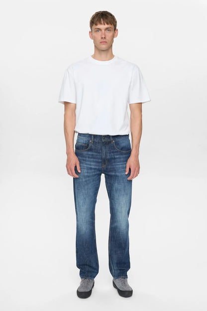 Vista laterale sinistra del modello che indossa i jeans Anerkjendt AKJUSTIN Dark Blue Sky con t-shirt bianca e sneaker grigie: profilo dei jeans straight fit che mostra la gamba dritta dal fianco all orlo grezzo con texture jacquard su denim blu medio su sfondo bianco