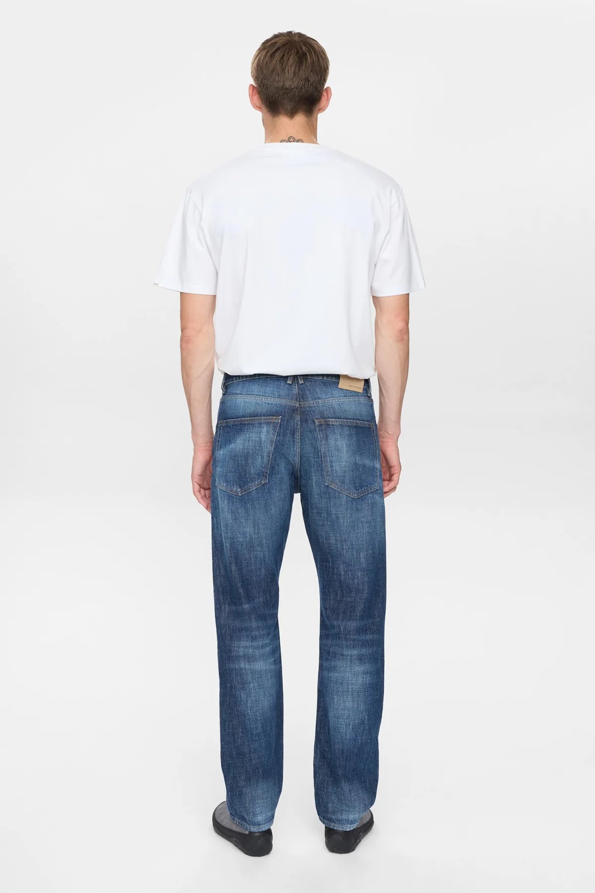 Vista laterale destra del modello che indossa i jeans Anerkjendt AKJUSTIN Dark Blue Sky con t-shirt bianca e sneaker grigie: profilo completo testa-piedi dei jeans straight fit con texture jacquard all-over lettera A su denim blu medio e orlo grezzo su sfondo bianco
