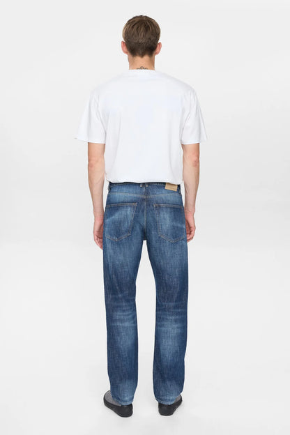 Vista laterale destra del modello che indossa i jeans Anerkjendt AKJUSTIN Dark Blue Sky con t-shirt bianca e sneaker grigie: profilo completo testa-piedi dei jeans straight fit con texture jacquard all-over lettera A su denim blu medio e orlo grezzo su sfondo bianco